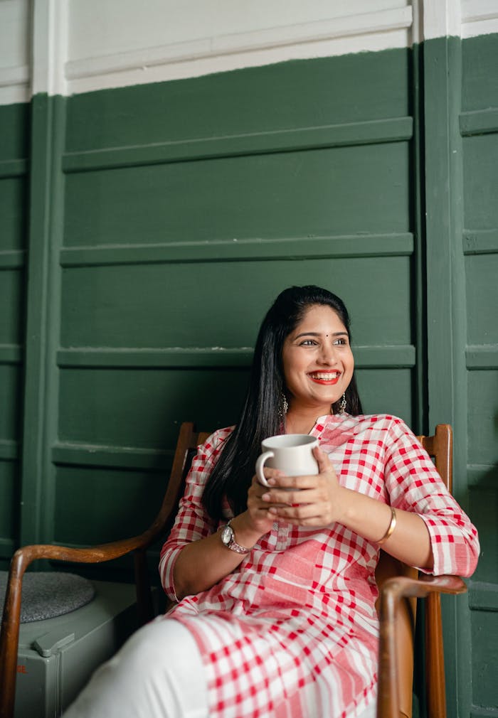 Positive charming young Indian lady in traditional pink and white wear resting on wooden chair with cup of hot beverage and looking away happily